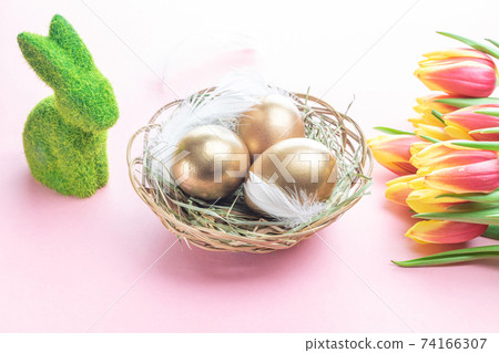 Egg color. Happy Easter decoration: golden colour eggs in basket with spring tulips, white feathers on pastel pink background. Foil minimalist egg design, modern template. 74166307