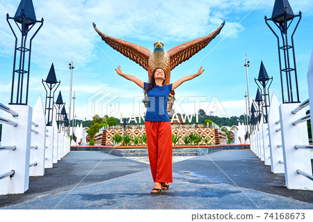 Happy girl tourist posing next to sculpture of a red eagle spreading its wings. Popular tourist spot on Langkawi island 74168673