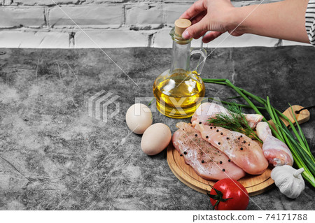 Woman holding a glass of oil and raw chicken fillet and legs on wooden plate with fresh vegetables Woman holding a glass of oil and raw chicken fillet and legs on wooden plate with fresh vegetables 74171788