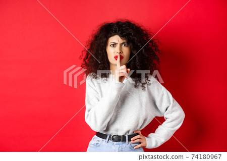 Angry young woman with curly hair and red lips, frowning and shushing, tell to be quiet, make taboo gesture to keep mouth shut, standing on red background Angry young woman with curly hair and red lips, frowning and shushing, tell to be quiet, make taboo gesture to keep mouth shut, standing on red background 74180967