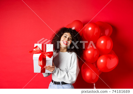 Holidays and celebration. Happy birthday girl holding gift and posing near party helium balloons, smiling excited at camera, red background Holidays and celebration. Happy birthday girl holding gift and posing near party helium balloons, smiling excited at camera, red background 74181689