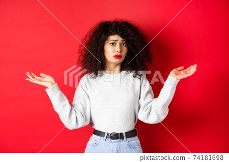 Who knows. Clueless young woman with curly hair, shrugging and looking confused, standing in casual clothes on red background 74181698