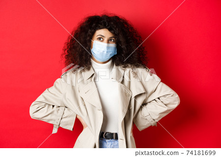 Covid-19, pandemic and quarantine concept. Stylish enthusiastic woman in medical mask and trench coat, smiling and looking at upper left corner, red background 74181699