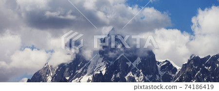 Panoramic view on high rocky mountains with snow and glacier hidden in fog and blue cloudy sky Panoramic view on high rocky mountains with snow and glacier hidden in fog and blue cloudy sky 74186419
