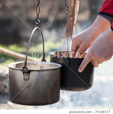 Man cooking in sooty old pots on bonfire Man cooking in sooty old pots on bonfire 74186577