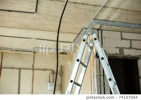 Aluminium ladder and electrical cables laid in protective corrugation installed on the ceiling and wall in a room under construction works. 74187084