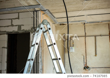 Aluminium ladder and electrical cables laid in protective corrugation installed on the ceiling and wall in a room under construction works. Aluminium ladder and electrical cables laid in protective corrugation installed on the ceiling and wall in a room under construction works. 74187085
