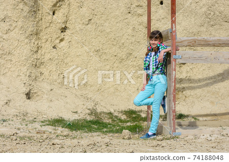 Stylish cute girl in a plaid shirt and jeans leans on a wooden structure against a background of a rock 74188048