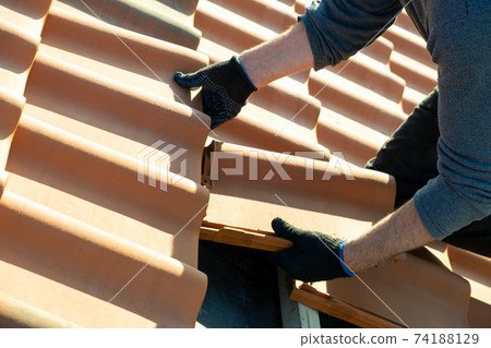Closeup of worker hands installing yellow ceramic roofing tiles mounted on wooden boards covering residential building roof under construction. 74188129