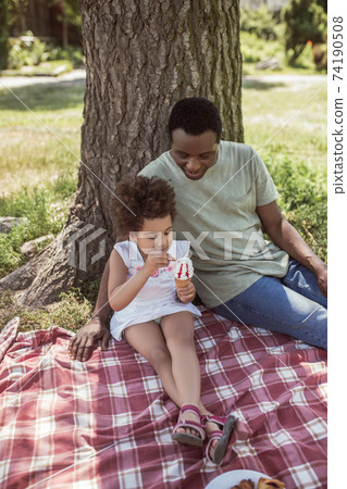 Dark-skinned cute kid sitting under the tree with dad and eating ice-cream Dark-skinned cute kid sitting under the tree with dad and eating ice-cream 74190508
