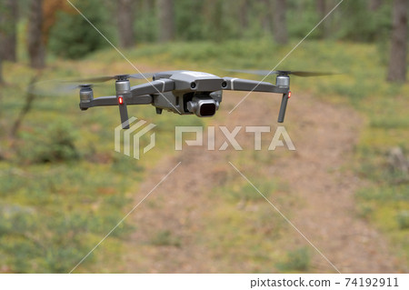 A drone quadrocopter with a camera with an ND filter hovers above the ground in flight and watches the road in the forest. Shallow depth of field, blurred background. 74192911