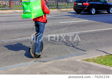 A modern concept of food delivery, the courier travels on an electric unicycle along the carriageway of a city street with a large green backpack on his back. 74201159