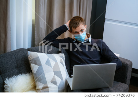 young man sitting on sofa with laptop in medical mask 74209376