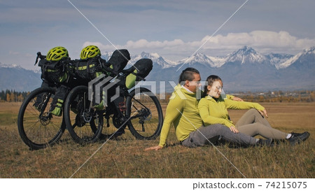 The man and woman travel on mixed terrain cycle touring with bikepacking. The two people journey with bicycle bags. Mountain snow capped. The man and woman travel on mixed terrain cycle touring with bikepacking. The two people journey with bicycle bags. Mountain snow capped. 74215075