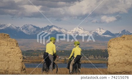 The man and woman travel on mixed terrain cycle touring with bikepacking. The two people journey with bicycle bags. Mountain snow capped, stone arch. The man and woman travel on mixed terrain cycle touring with bikepacking. The two people journey with bicycle bags. Mountain snow capped, stone arch. 74215076