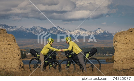 The man and woman travel on mixed terrain cycle touring with bikepacking. The two people journey with bicycle bags. Mountain snow capped, stone arch. The man and woman travel on mixed terrain cycle touring with bikepacking. The two people journey with bicycle bags. Mountain snow capped, stone arch. 74215077