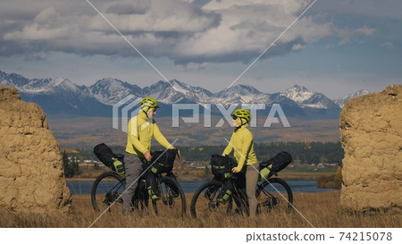 The man and woman travel on mixed terrain cycle touring with bikepacking. The two people journey with bicycle bags. Mountain snow capped, stone arch. The man and woman travel on mixed terrain cycle touring with bikepacking. The two people journey with bicycle bags. Mountain snow capped, stone arch. 74215078