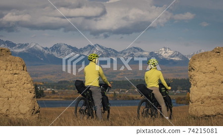 The man and woman travel on mixed terrain cycle touring with bikepacking. The two people journey with bicycle bags. Mountain snow capped, stone arch. The man and woman travel on mixed terrain cycle touring with bikepacking. The two people journey with bicycle bags. Mountain snow capped, stone arch. 74215081