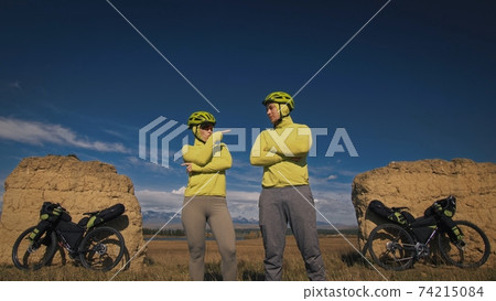 The man and woman travel on mixed terrain cycle touring with bikepacking. The two people journey with bicycle bags. Mountain snow capped, stone arch. The man and woman travel on mixed terrain cycle touring with bikepacking. The two people journey with bicycle bags. Mountain snow capped, stone arch. 74215084
