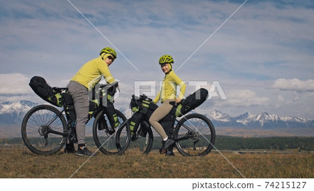 The man and woman travel on mixed terrain cycle touring with bikepacking. The two people journey with bicycle bags. Mountain snow capped. The man and woman travel on mixed terrain cycle touring with bikepacking. The two people journey with bicycle bags. Mountain snow capped. 74215127