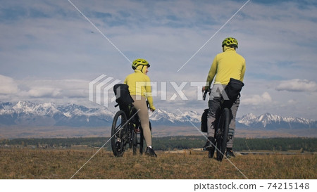 The man and woman travel on mixed terrain cycle touring with bikepacking. The two people journey with bicycle bags. Mountain snow capped. The man and woman travel on mixed terrain cycle touring with bikepacking. The two people journey with bicycle bags. Mountain snow capped. 74215148