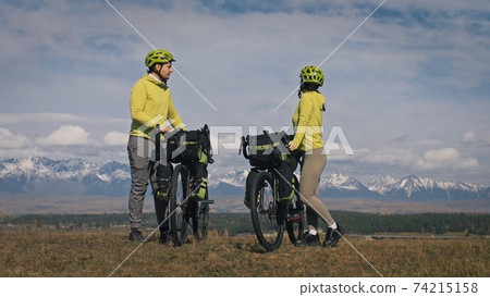 The man and woman travel on mixed terrain cycle touring with bikepacking. The two people journey with bicycle bags. Mountain snow capped. The man and woman travel on mixed terrain cycle touring with bikepacking. The two people journey with bicycle bags. Mountain snow capped. 74215158