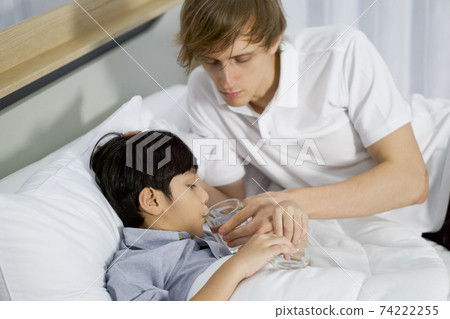 Young father takes care of his sick adopted son in bed. The boy tried to take medicine to improve the health condition. Young father takes care of his sick adopted son in bed. The boy tried to take medicine to improve the health condition. 74222255
