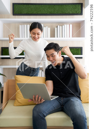 Young lovers spend time together on holidays in the living room. Both raised their fist up and smiled happily at the good news they received in e-mail while the man hold laptop computer. Young lovers spend time together on holidays in the living room. Both raised their fist up and smiled happily at the good news they received in e-mail while the man hold laptop computer. 74222596