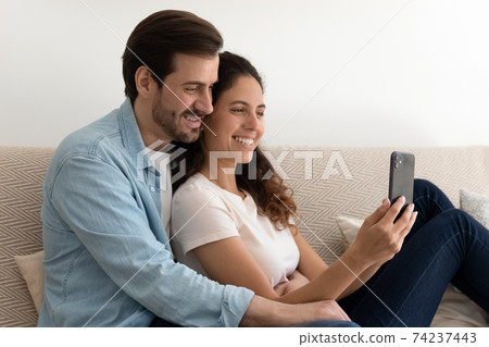 Positive married couple relaxing at home on couch holding cellphone 74237443