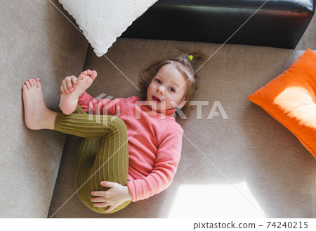a little girl in a pink sweatshirt is lying on the sofa among the pillows with her legs up. Home games. a little girl in a pink sweatshirt is lying on the sofa among the pillows with her legs up. Home games. 74240215