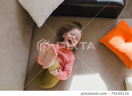 a little girl in a pink sweatshirt is lying on the sofa among the pillows with her legs up. Home games. 74240218