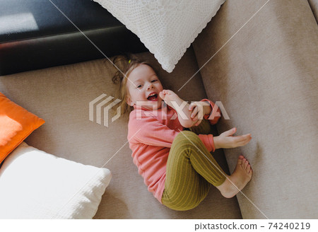 a little girl in a pink sweatshirt is lying on the sofa among the pillows with her legs up. Home games. a little girl in a pink sweatshirt is lying on the sofa among the pillows with her legs up. Home games. 74240219