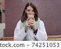 Smiling woman in winter coat with a coffee at the wall 74251053