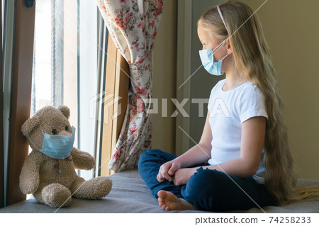 Stay at home quarantine coronavirus pandemic prevention. Sad girl and his teddy bear both in protective medical masks sitting next to the window and looks out window. Stay at home quarantine coronavirus pandemic prevention. Sad girl and his teddy bear both in protective medical masks sitting next to the window and looks out window. 74258233