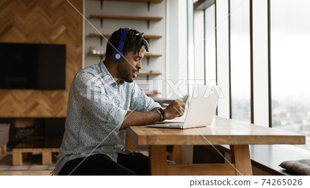 Young african guy use headphones laptop make notes of webinar 74265026
