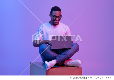 Black Programmer Guy Using Laptop Computer, Sitting On Cube In Neon Lighting 74269337