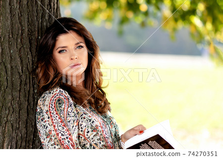Portrait of a sensual and lovely woman with nice hairstyle reading book outdoor on a sunny day under the tree ( copy space) 74270351