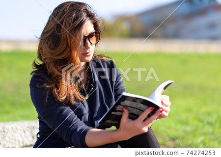 Beautiful young woman with a nice hairstyle and sunglasses sitting outdoor and reading a book on a sunny day. Beautiful young woman with a nice hairstyle and sunglasses sitting outdoor and reading a book on a sunny day. 74274253