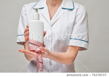 Portrait of beauty specialist standing against grey background and holding in her hand bottle of body lotion and measure tape 74276537