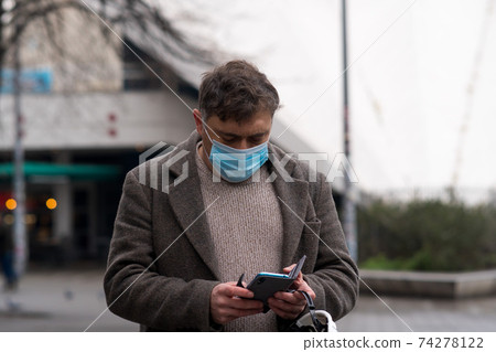 Outdoor portrait of a man covering his face with a medical mask to protect his nose and mouth looking down at his smartphone Outdoor portrait of a man covering his face with a medical mask to protect his nose and mouth looking down at his smartphone 74278122