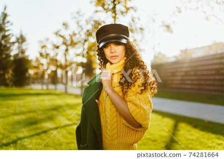 In the autumn park, a girl in a leather cap and a bright yellow sweater slung over her shoulder a green jacket with her hair with dark curls scattered over her shoulders In the autumn park, a girl in a leather cap and a bright yellow sweater slung over her shoulder a green jacket with her hair with dark curls scattered over her shoulders 74281664