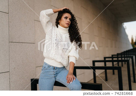 Beautifully posing photographer brunette in jeans and a long sweater, lifting his hand with his curly hair Beautifully posing photographer brunette in jeans and a long sweater, lifting his hand with his curly hair 74281668