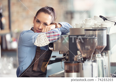 Bored waitress leans against the coffee machine with no job to do, holding face mask in her hand Bored waitress leans against the coffee machine with no job to do, holding face mask in her hand 74283203