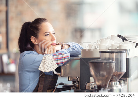 Bored waitress leans against the coffee machine with no job to do, holding face mask in her hand Bored waitress leans against the coffee machine with no job to do, holding face mask in her hand 74283204