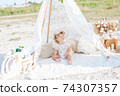 A little cute girl with red curly hair in summer, on the seashore in an airy lace bodysuit and a floral headband in a lace tent, devoried with marine details, shells and cotton. 1st BIRTHDAY.  74307357