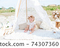 A little cute girl with red curly hair in summer, on the seashore in an airy lace bodysuit and a floral headband in a lace tent, devoried with marine details, shells and cotton. 1st BIRTHDAY.  74307360