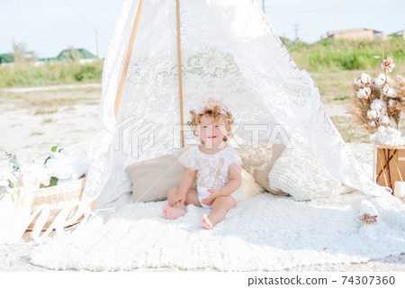 A little cute girl with red curly hair in summer, on the seashore in an airy lace bodysuit and a floral headband in a lace tent, devoried with marine details, shells and cotton. 1st BIRTHDAY.  74307360