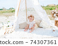 A little cute girl with red curly hair in summer, on the seashore in an airy lace bodysuit and a floral headband in a lace tent, devoried with marine details, shells and cotton. 1st BIRTHDAY.  74307361