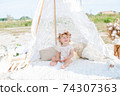 A little cute girl with red curly hair in summer, on the seashore in an airy lace bodysuit and a floral headband in a lace tent, devoried with marine details, shells and cotton. 1st BIRTHDAY.  74307363