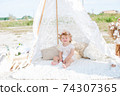 A little cute girl with red curly hair in summer, on the seashore in an airy lace bodysuit and a floral headband in a lace tent, devoried with marine details, shells and cotton. 1st BIRTHDAY.  74307365
